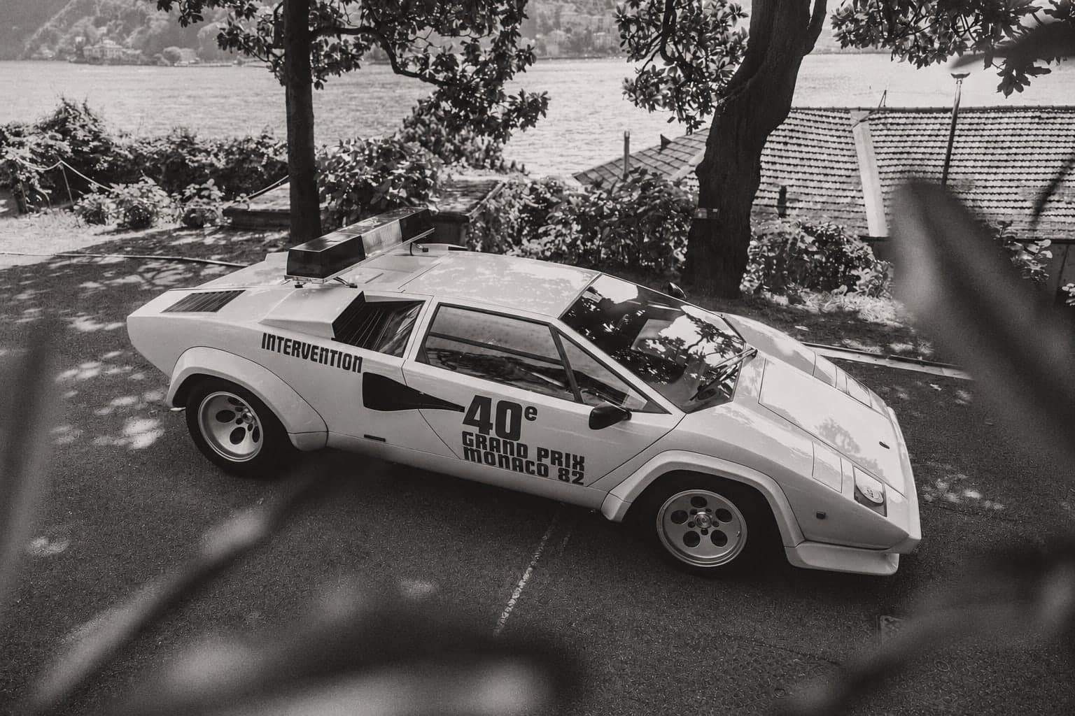 A black-and-white photograph of a white Lamborghini Countach with a roof light bar and "Intervention 40e Grand Prix Monaco 82" livery, parked beneath trees beside a lake.