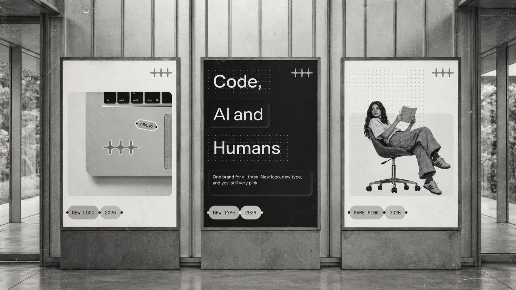 Three posters in n8n's new brand identity on display stands, one with a laptop and an n8n sticker, one reading "Code, AI and Humans", and one with a woman reading in an office chair.