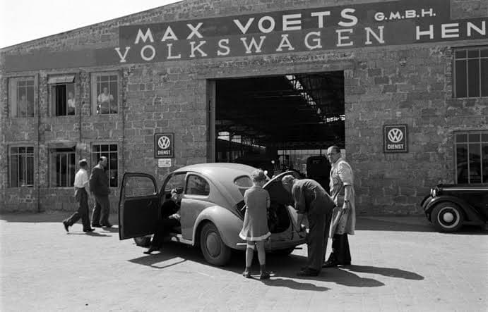 A black-and-white photograph of a Volkswagen Beetle with its door and rear engine lid open outside the Max Voets G.m.b.H. Volkswagen dealership, as people look on.